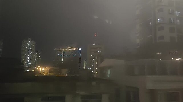 Nighttime cityscape view of illuminated Bangkok skyscrapers and buildings captured through a BTS Skytrain window.