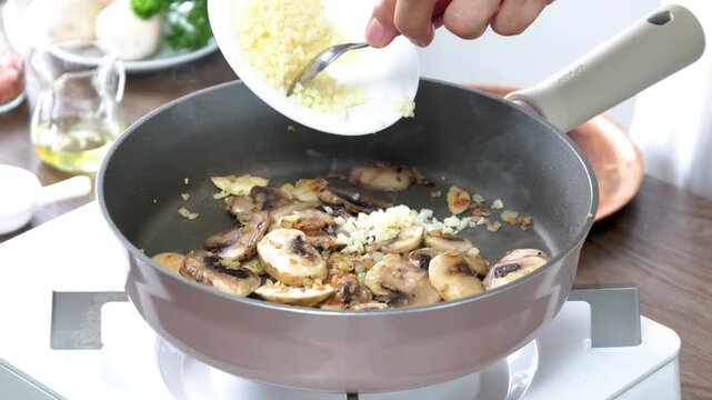 Adding freshly minced garlic to saut&eacute;ed mushrooms in a hot skillet, creating a rich aroma and flavorful base for homemade cooking. Close-up of a chef preparing a healthy meal in a modern kitchen with 