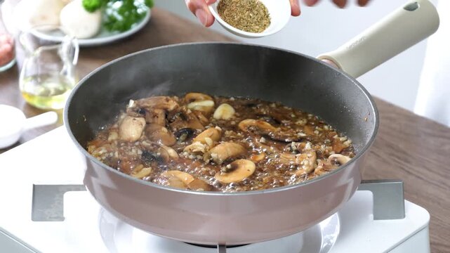 Hand sprinkling dried herbs Oregano into a creamy mushroom sauce simmering in a frying pan. Homemade cooking with fresh mushrooms, garlic, and aromatic seasonings on a stovetop. Close-up food preparat