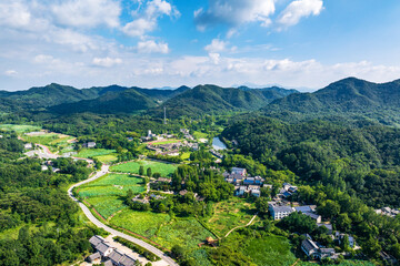 Obraz premium An aerial shot of a rural village nestled among lush green mountains, farmlands and forests under a bright blue sky with white clouds.