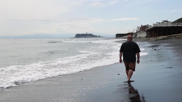 Man walking alone on a gray sandy beach in Kamakura, Japan, ocean waves rolling in and Enoshima island in the background. Moody coastal travel atmosphere.