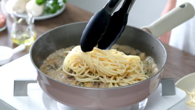 Fresh spaghetti being added to a creamy mushroom sauce in a frying pan during home cooking. Close-up of a delicious Italian pasta recipe in progress, featuring boiling cream sauce, mushrooms, and perf