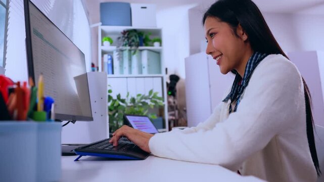 Asian woman typing on keyboard at office desk with computer monitor, plant, stationery, modern workspace, focused indoor worker using desktop screen in professional workplace