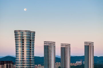 Several modern high-rise towers stand against a twilight sky with a visible moon and distant hills. © tai
