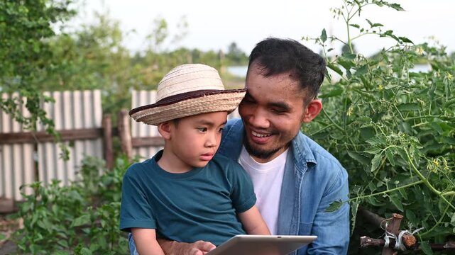 Modern Asian farmer teaching young son agricultural technology using digital tablet in organic garden