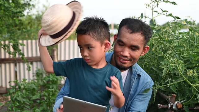 Modern Asian farmer teaching young son agricultural technology using digital tablet in organic garden