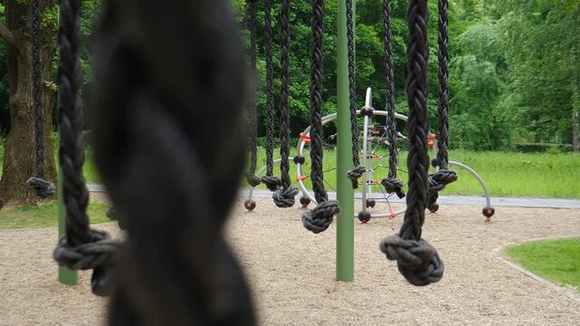 Outdoor city park playground with hanging braided ropes and green metal posts. Blurred child running in the background, framed by a large out-of-focus rope in the foreground 4k video