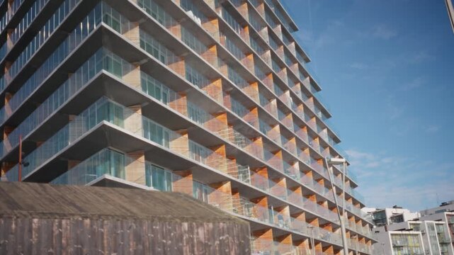 A contemporary apartment building with rows of glass balconies and wooden slat screens stands beneath a bright blue sky, wide shot. Aarhus, Denmark