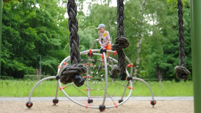 Close-up of a metal spherical climbing frame with thick ropes and red-tipped legs. A blurred child plays in the lush green city park background 4k video