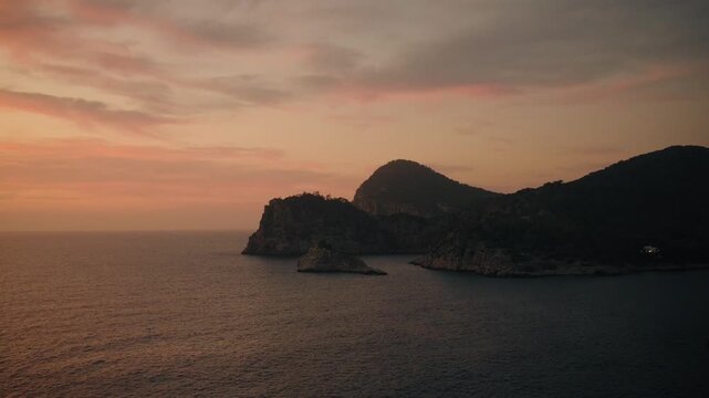 Sweeping aerial footage captures Es Vedr&aacute; island and Cala d'Hort&rsquo;s rugged coastline at golden hour, with tranquil waters, dramatic silhouettes, and vibrant, colorful sunset skies.