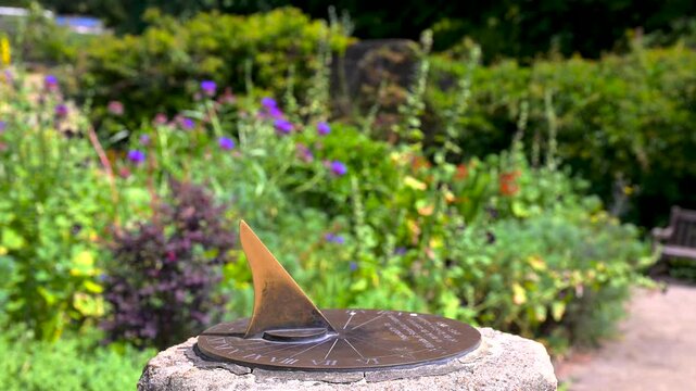 Roman sundial on a stone plinth in a soft-focus flower border on a still, bright day.
