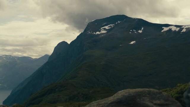 Steep mountain ridge rises above a deep valley with scattered summer snowfields under a heavy sky.