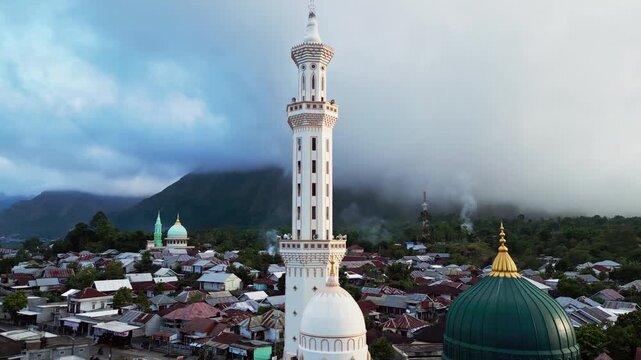 Islamic mosque with a green dome and towering minaret surrounded by village homes beneath cloud covered volcanic mountains, aerial drone footage in Indonesia.