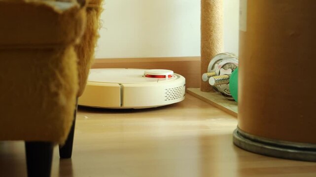 Horizontal 4K video showing a white smart robot vacuum cleaner navigating and cleaning dust under a chair on parquet wooden flooring