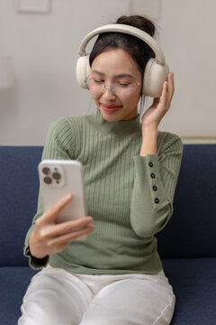 Happy young Asian woman relaxing on comfortable blue sofa holding modern smartphone wearing wireless headphone listening to favorite music feeling joyful peaceful indoor living room space