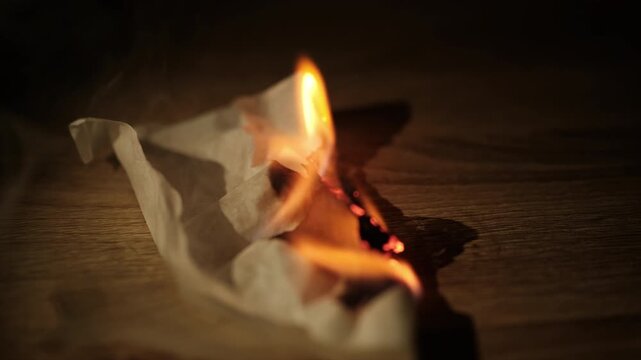 Close-up of burning paper tissue with bright flame on a wooden table in dark low light