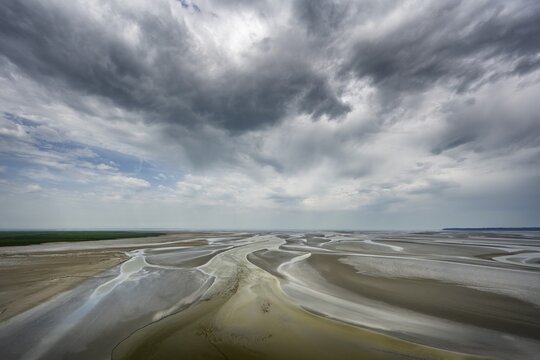 View from the rocky island, monastery island of Le Mont Saint Michel onto a wide mudflat landscape with creeks and dramatic clouds at low tide, Mont-Saint-Michel Bay, Pontorson, Manche department, Normandy, France