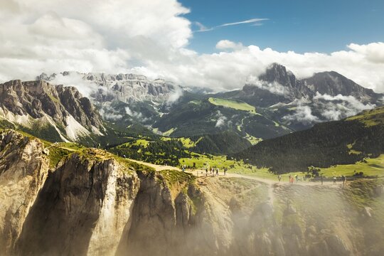 Aerial panorama tourist walk hike Seceda ridge with Odle peaks rise over clouds with steep limestone cliffs green alpine slopes winding hiking trail. Panoramic Dolomites landscape. South Tyrol Italy