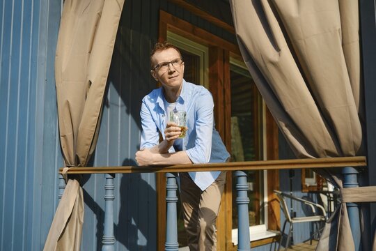 Middle-aged man in blue shirt and beige trousers stands on terrace of wooden cabin looking thoughtful in distance while holding glass in his hand