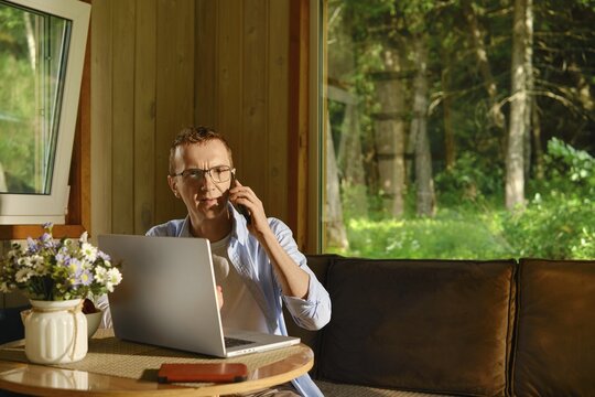 Middle-aged man sits at table in wooden cabin, talking on phone while using laptop. He solves problems over the phone remotely. Green trees visible through windows. Sunlight shines inside creating bright atmosphere during daytime