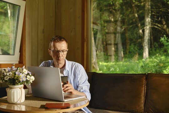 Middle-aged man works remotely from wooden country cabin. There is laptop on table in front of him and he is typing text message on his smartphone. Forest is visible through large windows