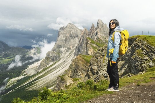 Happy female traveler yellow backpack stand on famous viewpoint ridge line Seceda South Tyrol Dolomites Italy looking up feeling free peaceful active lifestyle outdoor vacation