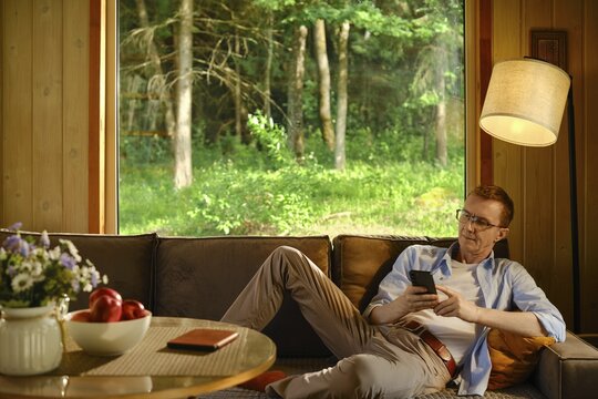 Middle-aged man rests on sofa inside wooden cabin with air conditioner and looks at smartphone under floor lamp. Concept of quiet and relaxing time outside city in nature