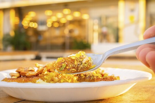 Jordan kunafa dessert close up in paper plate with sugar syrup melted cheese pistachio cashew topping spoon bokeh cafe background