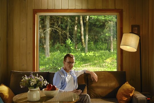Middle-aged man hiding from heat in wooden cabin outside city sitting on couch and reading electronic book. Sunlight enters through large window. Outside, there are trees and green grass