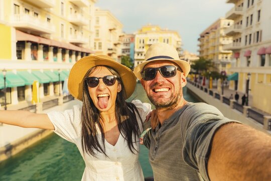 Happy tourist couple taking selfie Qanat Quartier Doha. Woman sticking tongue out, man smiling, straw hats, sunglasses. Venetian style canals, colorful buildings background