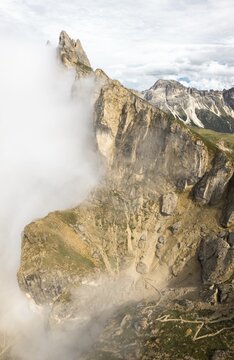 Dramatic drone aerial view jagged rock cliffs Seceda ridge line partly covered rolling white clouds switchback hiking trail valley alpine landscape South Tyrol Dolomites Italy