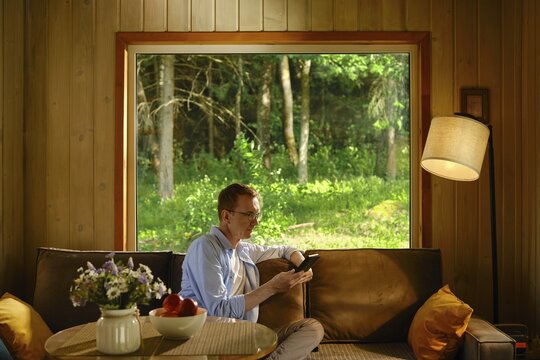 Middle-aged man sits on couch in wooden cabin, holding electronic book and reading a novel. Light streams through large window. Green trees show outside