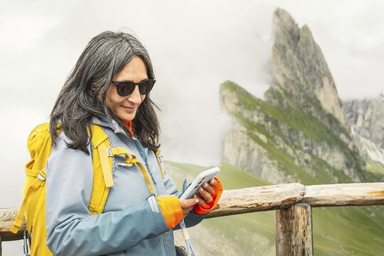Smiling woman hiker checking mobile GPS navigation app type message, use social media on smartphone using satellite connectivity or esim in Seceda mountain ridge walk. Dolomites Italy holidays