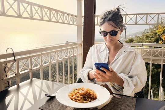 Woman taking photo eating shrimp spaghetti on scenic Mediterranean restaurant terrace. Golden hour lighting, coastline views. Delicious seafood healthy fresh traditional eating on summer holiday