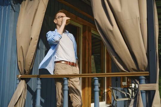 Middle-aged man stands at railing of patio terrace enjoying cold drink under hot afternoon sun