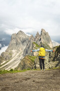 Back view middle aged gray hair woman hiker arms outstretched with yellow backpack overlooking lush green alpine valley misty mountains background Dolomites Alps Italy. Seceda ridge viewpoint