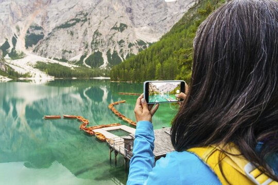 Woman hiker taking smartphone landscape photo of Lago di Braies Lake in italian Dolomites mountains in northern Italy. Drone aerial photo with Wooden boats and beautiful reflection in calm water