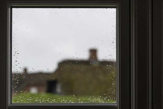 View through a window pane with raindrops beading, blurred traditional thatched Danish houses in the background, Houvig, North Sea, Denmark