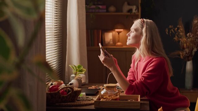 Young adult Caucasian woman sitting at table, listening to music on smartphone with earphones, smiling and looking out window. Clip is suitable for mental health apps or audio streaming ads