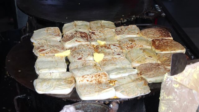 A folded Martabak Telor Kubang pastry filled with seasoned eggs, minced beef, onions, and aromatic spices is carefully placed onto a large hot griddle for cooking. 