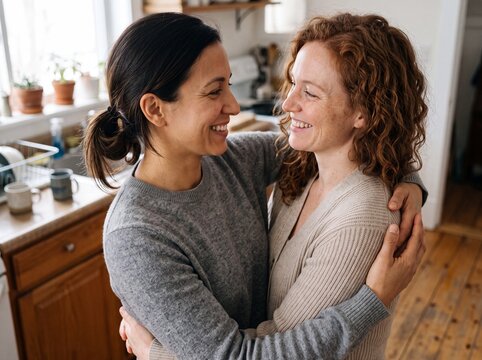 Zwei l&auml;chelnde Frauen in ihren Drei&szlig;igern umarmen sich liebevoll mit Blickkontakt in einer hellen wohnlichen K&uuml;che
