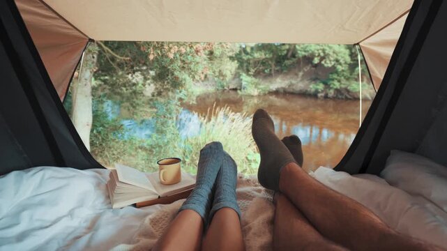Feet wearing cozy warm socks rest on a bed inside a camping tent looking out at a calm river scene while an open book and a metal mug sit nearby to create a peaceful atmosphere for relaxing time.