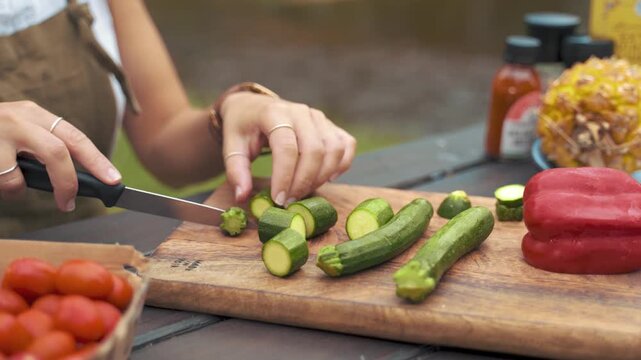 Zucchini vegetables sit on a rustic wooden cutting board while a pair of hands carefully slices the fresh produce with a sharp silver knife in an outdoor setting filled with healthy natural.