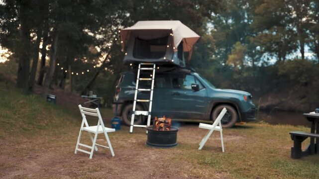 Vehicle featuring a mounted roof tent stands parked in a serene forest clearing with empty folding chairs placed near a fire pit ready for guests to arrive for an evening of relaxation and comfort.