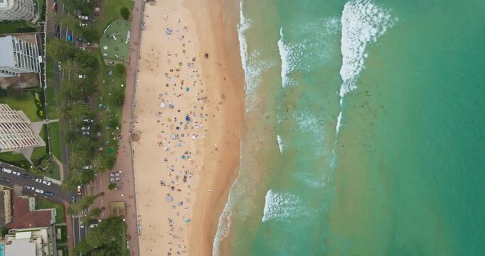 People are enjoying a summer relax day on Manly Beach in Sydney. Australia. Sand beach, clear turquoise water, ocean waves and road. Travel and vacation background. Aerial top down zoom in shot