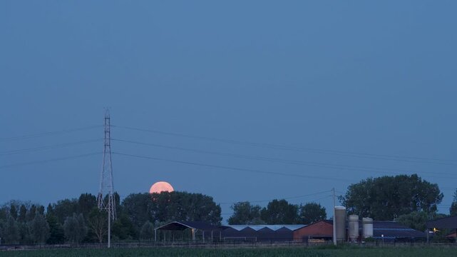 Strawberry Moon behind a scenic countryside farm at dusk with a rising full moon in a clear blue sky Tranquil rural landscape atmospheric lighting vibrant colors smooth motion