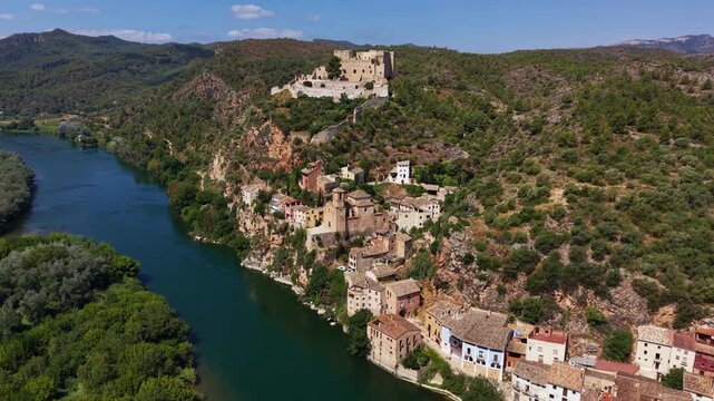 Kayakers in yellow boats paddle along the Ebro River beside Miravet's historic cliffside village in Catalonia, Spain, with a medieval castle crowning the hilltop