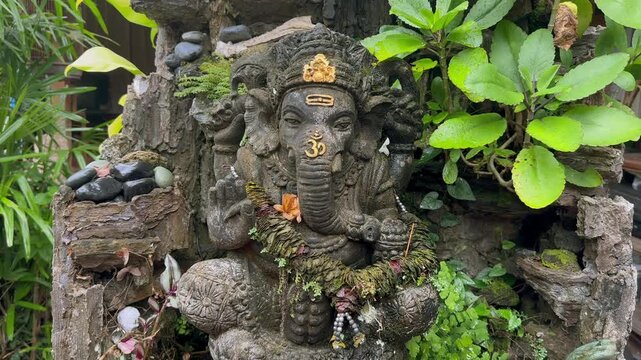 Stone sculpture of Ganesha stands among green plants beside an old stone wall in an outdoor temple setting. Religious symbolism.
