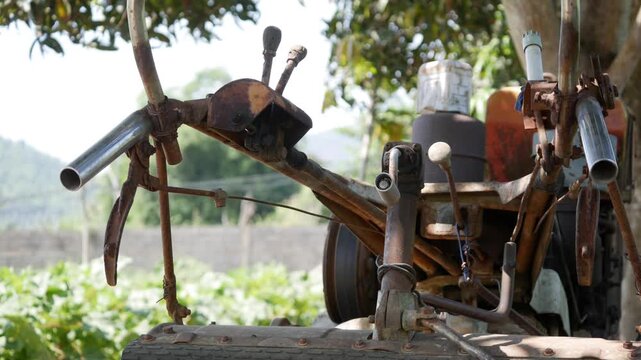 An old rusty small tractor with metal attachments stands in a green field with trees and open countryside behind it. Heritage implement.