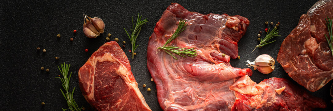 Different cuts of beef steak are arranged on a black surface. Fresh herbs, spices, and garlic accompany the meat, ready for cooking or grilling at a kitchen or market, banner